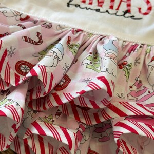 May include: A close-up of a white and pink Christmas-themed apron. The apron features a ruffled skirt with a candy cane stripe pattern and a repeating pattern of Santa, Christmas trees, and ornaments. The text "SANTA'S favorite" is embroidered on the bib.
