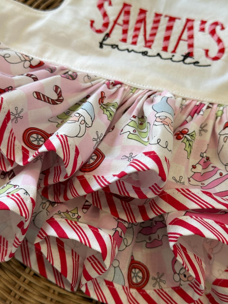 May include: A close-up of a white and pink Christmas-themed apron. The apron features a ruffled skirt with a candy cane stripe pattern and a repeating pattern of Santa, Christmas trees, and ornaments. The text "SANTA'S favorite" is embroidered on the bib.