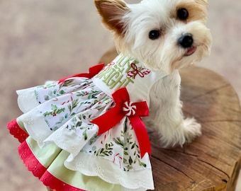 May include: A small dog wearing a white sleeveless dress with a tiered skirt featuring a green and red holiday botanical print. The bodice has green embroidered text reading "LUCK BRIGHT" and a red satin bow with a peppermint candy accent.
