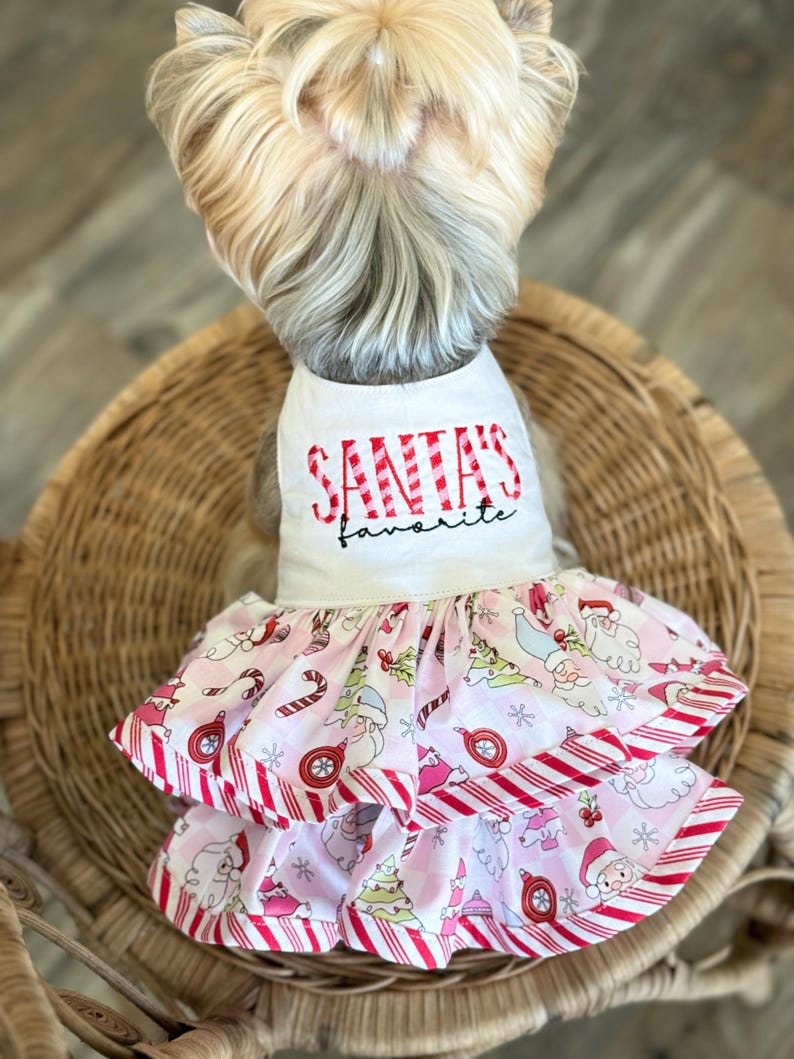 May include: A dog wearing a white and pink Christmas dress. The dress has the text "SANTA'S favorite" in red. The skirt of the dress features a pattern of Santa Claus, Christmas trees, and candy canes. The dress has red and white striped trim.