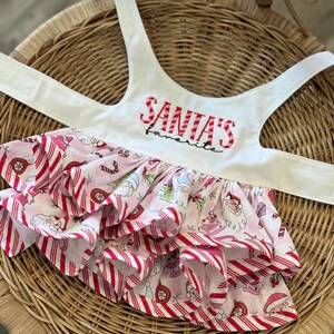May include: A white pet harness with the text "SANTA'S favorite" in red embroidery. The skirt is pink with a candy cane stripe pattern and a repeating Christmas-themed print, including Santa Claus and ornaments.