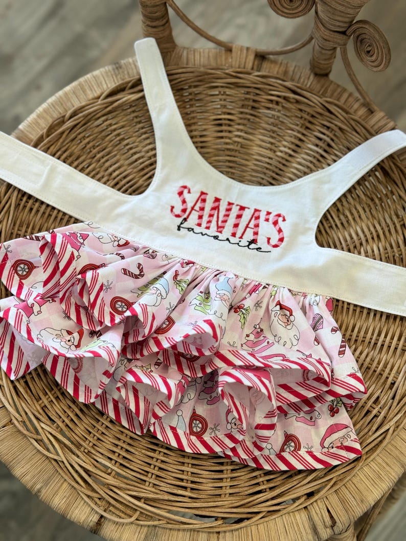 May include: A white pet harness with the text "SANTA'S favorite" in red embroidery. The skirt is pink with a candy cane stripe pattern and a repeating Christmas-themed print, including Santa Claus and ornaments.