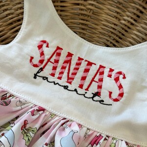 May include: A white and pink Christmas dress with the text "SANTA'S favorite" embroidered in red. The dress features a pink and white patterned skirt with Christmas gnome and holly illustrations.