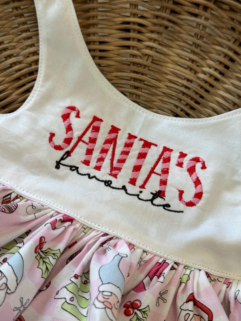 May include: A white and pink Christmas dress with the text "SANTA'S favorite" embroidered in red. The dress features a pink and white patterned skirt with Christmas gnome and holly illustrations.