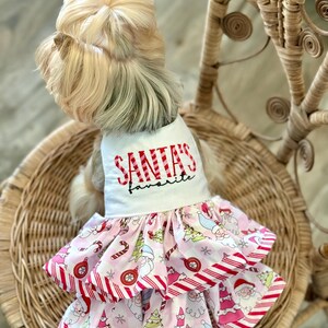 May include: A small dog wearing a white and pink Christmas dress. The dress has a white top with the text "SANTA'S favorite" in red script. The skirt is pink with a candy cane and Santa Claus pattern.