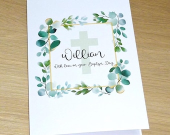 May include: A white baptism card featuring a gold-framed cross and the name "William" in cursive. The card includes the text "With love on your Baptism Day" and is adorned with green and gold eucalyptus leaves, creating a delicate, celebratory design.