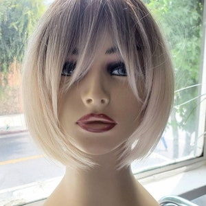 Puede incluir: Una peluca rubia corta con raya lateral y flequillo. La peluca está peinada con un corte recto y contundente con una ligera onda. La peluca está en una cabeza de maniquí.