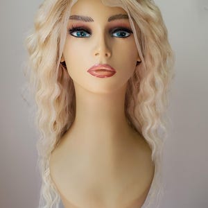 Op de afbeelding: Een blonde pruik met losse, golvende krullen. De pruik zit op een mannequinhoofd en heeft een natuurlijk ogende haarlijn.