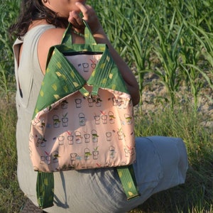 Puede incluir: Una mochila verde y rosa con un patrón de pequeños cubos y plantas. La mochila tiene correas ajustables y un cierre de cremallera.