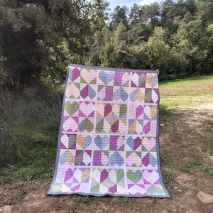 Puede incluir: Un edredón hecho a mano con un diseño de patchwork con forma de corazón en varios colores, incluyendo rosa, morado, verde y azul. El edredón tiene un borde azul claro y se muestra al aire libre.