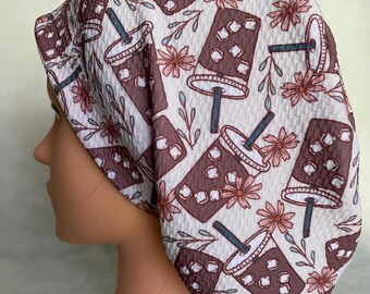 May include: A patterned scrub cap featuring a repeating design of iced coffee cups, pink flowers, and green leaves on a white background. The cap is made of a textured fabric and is designed to fit comfortably.