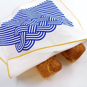 May include: A white fabric bread bag with a blue and yellow geometric pattern. The bag is folded over two loaves of bread.