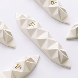 May include: Geometric white mezuzah cases with gold-colored Hebrew letters. The cases have a faceted design and are arranged on a white background. The cases are a religious item.