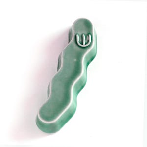 May include: A sage green ceramic chopstick rest with a wavy, elongated form and a smooth, glossy finish. The top surface features a small, embossed white symbol resembling a stylized trident or a three-pronged fork, centered near the rounded end.