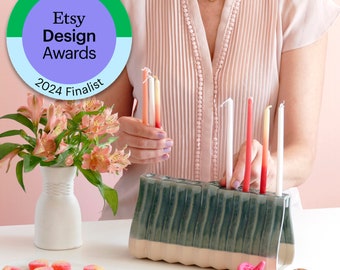 May include: A ceramic menorah with a green and cream design, holding lit and unlit candles. The image includes a vase of pink flowers, a plate of candies, and a pink dreidel. The Etsy Design Awards 2024 Finalist badge is in the upper left.