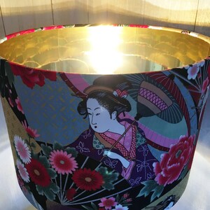 Puede incluir: Una pantalla de l&aacute;mpara con la parte superior dorada y la inferior de tela colorida. La tela presenta una geisha con un paraguas rosa y negro, flores y mariposas.