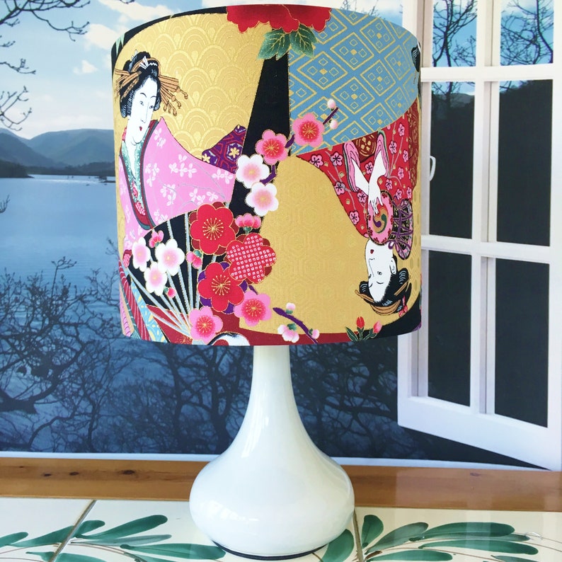 Puede incluir: Una l&aacute;mpara de mesa blanca con una pantalla cil&iacute;ndrica con un dise&ntilde;o de inspiraci&oacute;n japonesa. La pantalla est&aacute; decorada con un fondo dorado, flores de cerezo y dos mujeres con ropa tradicional japonesa.