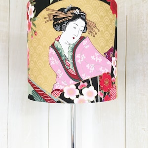 Puede incluir: Una pantalla de l&aacute;mpara con fondo negro y un dise&ntilde;o japon&eacute;s colorido con una mujer en un kimono rosa y flores de cerezo.