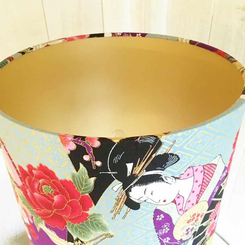 Puede incluir: Una pantalla de l&aacute;mpara con fondo azul y dorado que presenta un dise&ntilde;o de geisha japonesa con flores rosas y rojas. La geisha lleva un kimono rosa y blanco y sostiene un abanico.