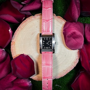 May include: A pink watch with a rectangular black face and silver-colored trim. The watch has a pink, textured leather band. The watch is displayed on a wooden slice, surrounded by dark red rose petals.