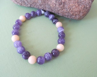 May include: A purple and cream beaded bracelet with a knotted closure. The bracelet is made with round beads of amethyst and cream colored stone.