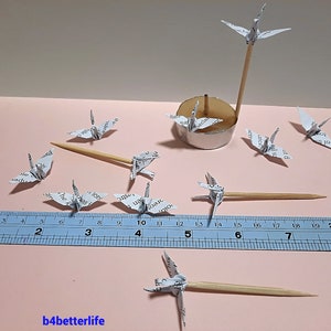 May include: A collection of white origami paper cranes, some perched on wooden skewers, others on a silver stand. A ruler is included for scale, measuring up to 8 inches.