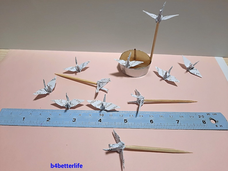 May include: A collection of white origami paper cranes, some perched on wooden skewers, others on a silver stand. A ruler is included for scale, measuring up to 8 inches.