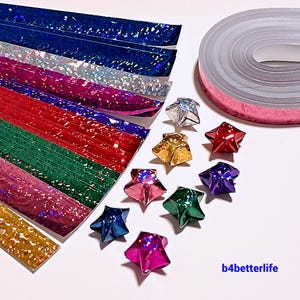 Op de afbeelding: Een assortiment van iriserende papieren stroken in verschillende kleuren, waaronder goud, groen, rood, roze, blauw en zilver. De stroken zijn in een waaier vorm gerangschikt. Er zijn ook verschillende origami sterren gemaakt van de papieren stroken in verschillende kleuren, waaronder goud, zilver, rood, roze, blauw en paars. De afbeelding bevat ook een rol roze iriserend papier.
