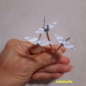 May include: Three paper origami cranes on wooden skewers. The cranes are made from white paper with black text. The text on the cranes includes the words "time", "with", "his", "even", "the", and "pro".