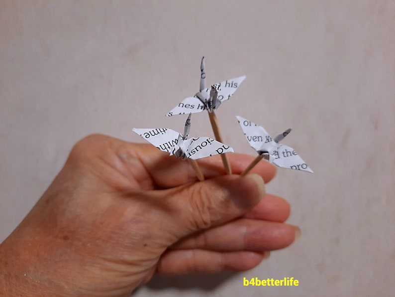 May include: Three paper origami cranes on wooden skewers. The cranes are made from white paper with black text. The text on the cranes includes the words "time", "with", "his", "even", "the", and "pro".
