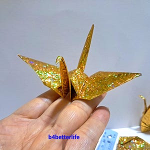 May include: A gold origami crane with glittery details is held in a hand. The crane is in flight, with its wings spread. The text "b4betterlife" is visible in the lower left corner. Other origami cranes are in the background.