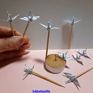 May include: A collection of paper cranes made from printed paper, each attached to a wooden skewer. The cranes are arranged around a small candle holder.