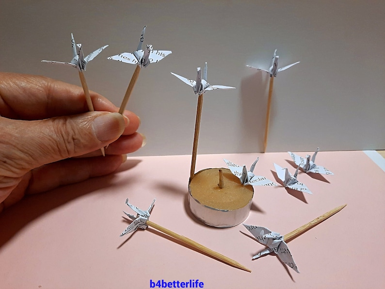 May include: A collection of paper cranes made from printed paper, each attached to a wooden skewer. The cranes are arranged around a small candle holder.