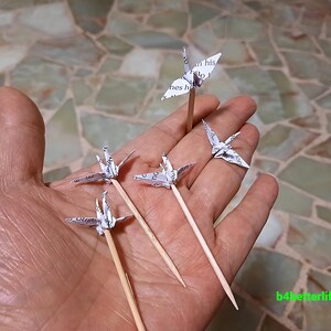 May include: Five white paper origami cranes on wooden skewers. The cranes are made from printed paper and are arranged in a hand.