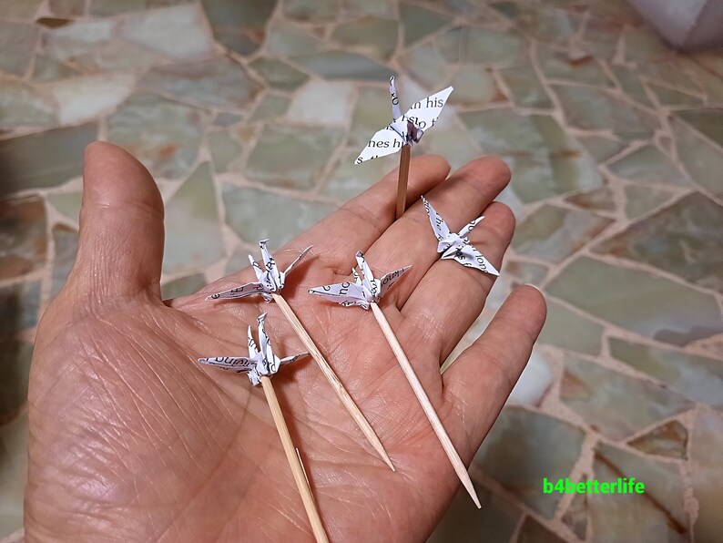May include: Five white paper origami cranes on wooden skewers. The cranes are made from printed paper and are arranged in a hand.
