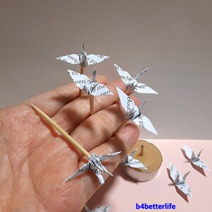 May include: A collection of white paper origami cranes, some on wooden skewers, with text printed on the paper. The cranes are arranged on a hand and a candle holder.
