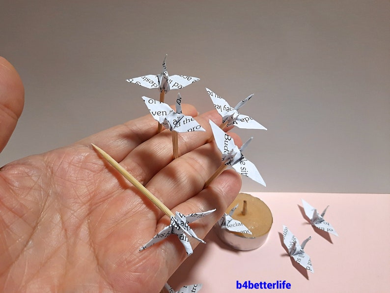 May include: A collection of white paper origami cranes, some on wooden skewers, with text printed on the paper. The cranes are arranged on a hand and a candle holder.