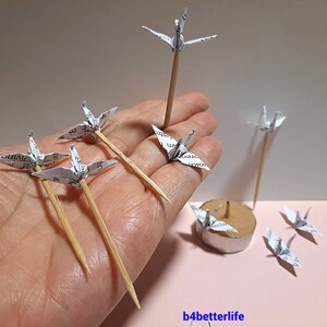May include: A collection of white paper origami cranes on wooden skewers. The cranes are arranged in a hand and on a small round base. The cranes are made from paper with printed text.