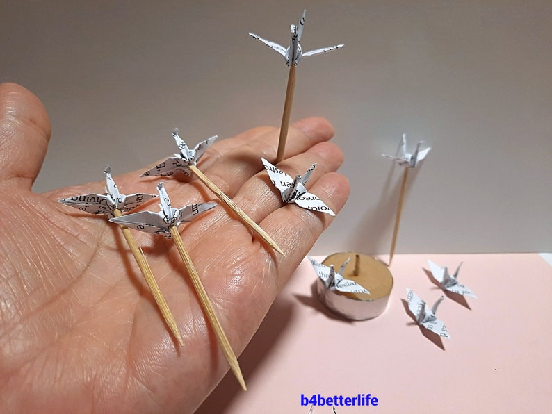 May include: A collection of white paper origami cranes on wooden skewers. The cranes are arranged in a hand and on a small round base. The cranes are made from paper with printed text.