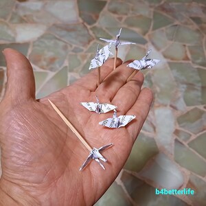 May include: Five white paper origami cranes on wooden skewers, held in a hand. The cranes are made from printed paper with text visible.