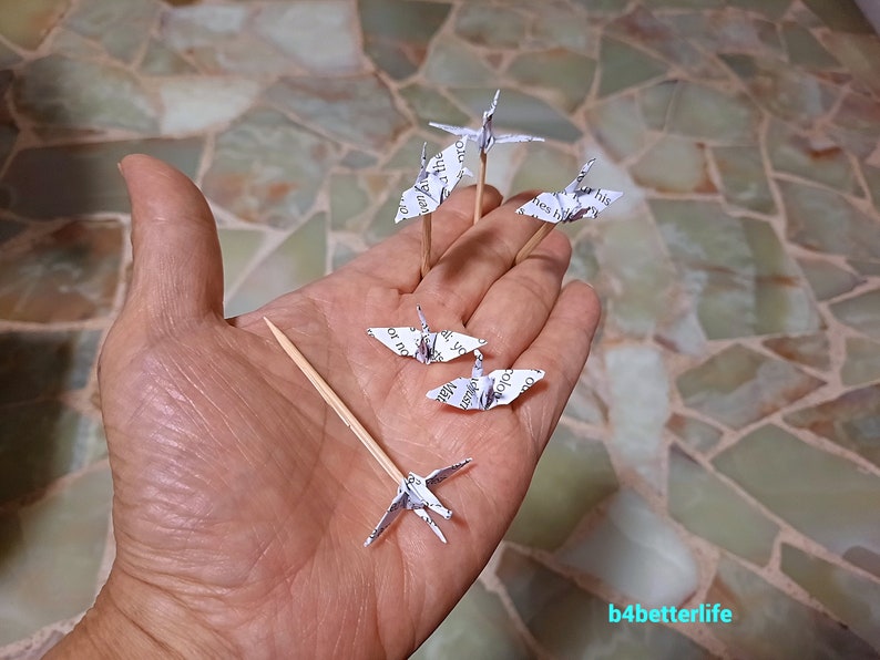May include: Five white paper origami cranes on wooden skewers, held in a hand. The cranes are made from printed paper with text visible.