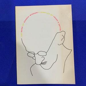 May include: A white book cover with a black line drawing of a person wearing glasses. The text "THE BOOK OF GANDHI WISDOM - COMPILED BY TRUDY S. SETTEL" is written in a semi-circle around the drawing.