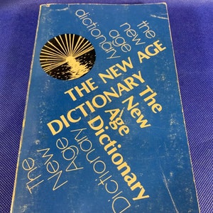 Könnte beinhalten: Ein blaues Buch mit dem Titel "The New Age Dictionary" in gelber Schrift. Der Buchumschlag zeigt eine Schwarzweißillustration eines Sonnenaufgangs über einem Gewässer.