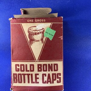 Könnte beinhalten: Eine Vintage-Box mit "GOLD BOND BOTTLE CAPS" in Rot und Creme. Die Box zeigt eine Zeichnung eines Flaschenverschlusses und die Worte "ONE GROSS". Auf der Seite befindet sich ein grüner Aufkleber.