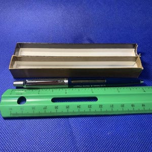 May include: A blue pen with silver accents and the text "U.S. News & World Report" is next to a green ruler. The pen is in front of a brown cardboard box. The ruler measures up to 30 inches.