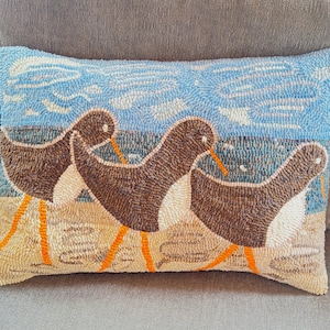 May include: Rectangular decorative pillow featuring a beach scene with three brown and white birds with orange legs and beaks. The background shows a blue sky and ocean. The pillow is made of a textured material.