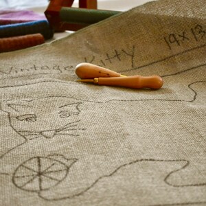 以下が含まれることがあります： ベージュのキャンバスに、黒い線で描かれた糸巻きを持った猫の絵が描かれています。キャンバスには「Vintage Kitty 19x13.5」とラベルが貼られています。キャンバスの上に、木製の針工具が2つ置かれています。