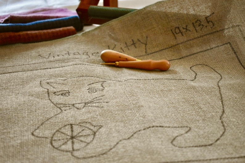 以下が含まれることがあります： ベージュのキャンバスに、黒い線で描かれた糸巻きを持った猫の絵が描かれています。キャンバスには「Vintage Kitty 19x13.5」とラベルが貼られています。キャンバスの上に、木製の針工具が2つ置かれています。