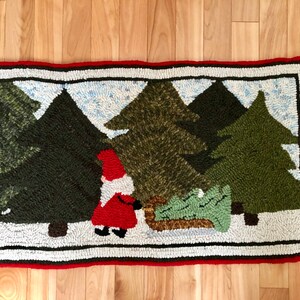 May include: A rectangular hooked rug featuring a scene of a gnome in a red coat and hat pulling a small green Christmas tree on a sled through a snowy forest of dark green pine trees, bordered by a thin black line and a thick red outer edge.