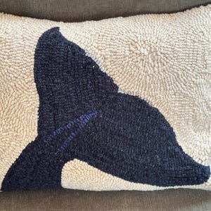May include: A rectangular decorative pillow with a cream-colored background and a navy blue whale tail design. The pillow has a textured, looped surface, and the whale tail is detailed with blue stitching.